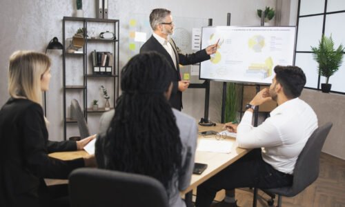 Male mature manager in business suit, speaking at office meeting with his multiethnic male and female colleagues, sitting at table, showing company's annual growth report charts and map on TV screen.